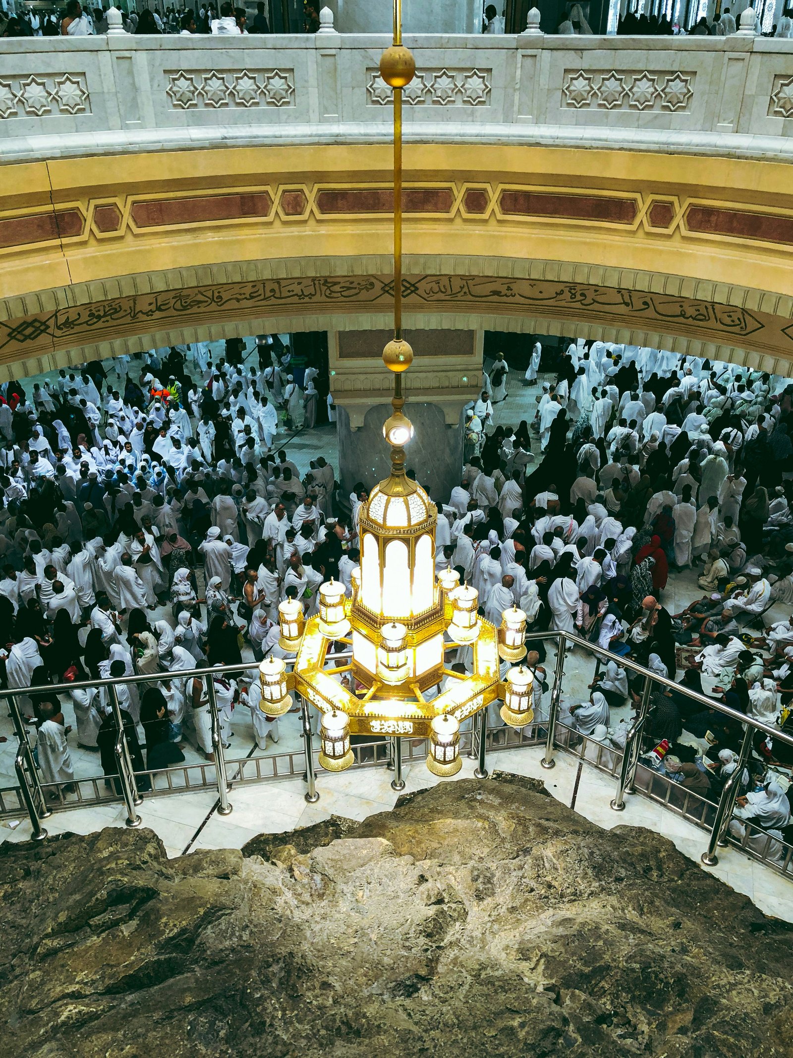 Overhead view of a large crowd of pilgrims dressed mostly in white gathered inside a mosque around a large rock, beneath a prominent ornate chandelier hanging from an arched ceiling with Arabic calligraphy.